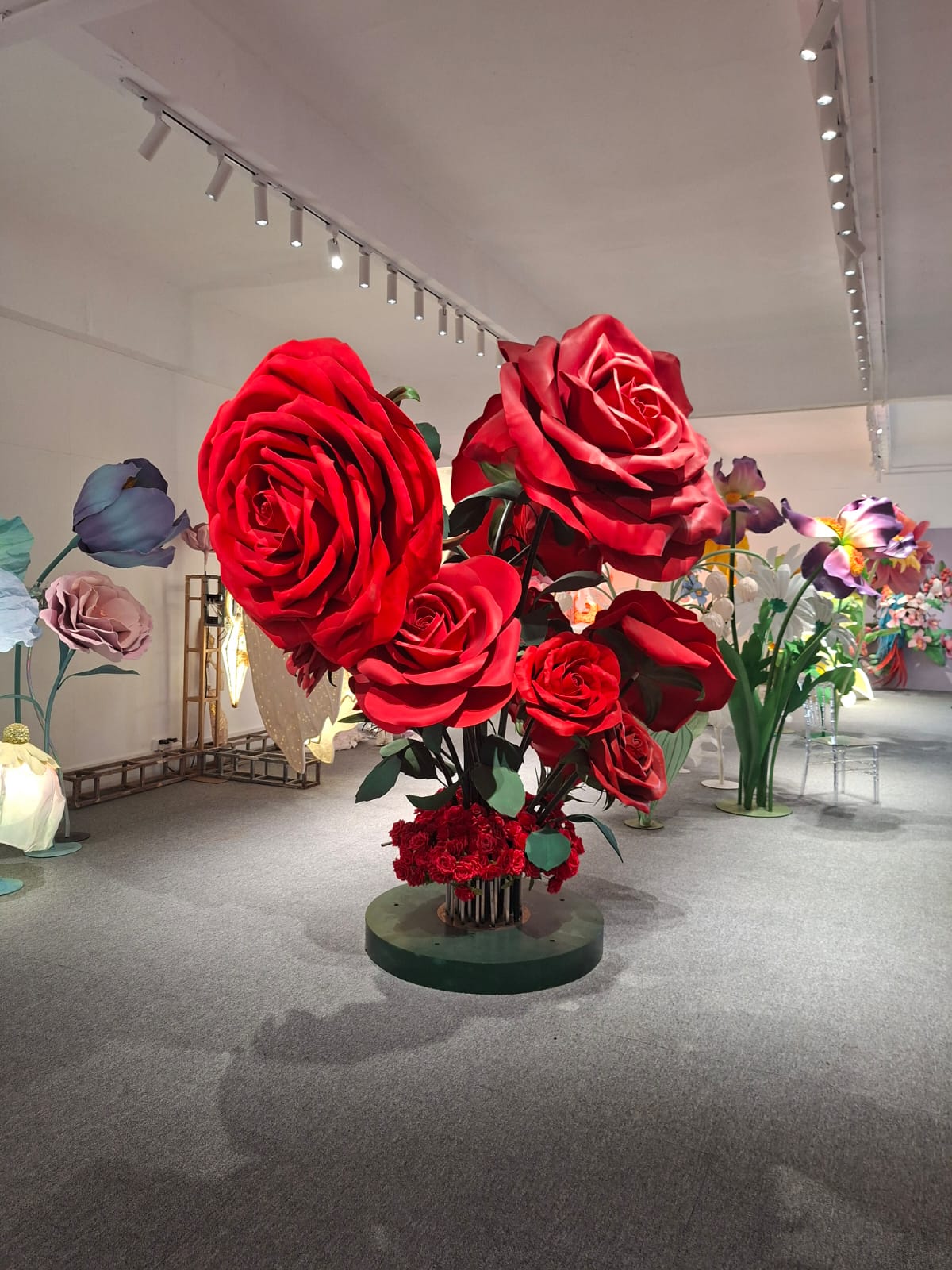 A towering bouquet of oversized red roses on a green pedestal inside the Sparking Joy studio, surrounded by other giant blooms